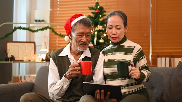 Image of happy senior couple using digital tablet on couch at home with Christmas tree behind.