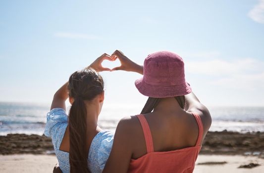 Beach, heart sign and women friends with love for summer and ocean on blue sky and sunshine. Behind of lesbian couple or gen z people with care hands, icon or emoji for outdoor vitamin d or earth day.
