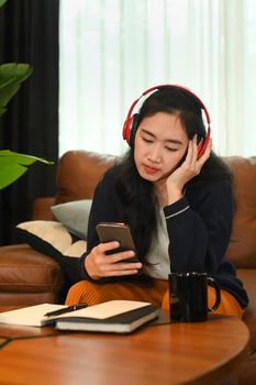 Pretty young asian woman wearing wireless headphone and using mobile phone on couch at home.