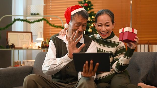 Affectionate senior couple making video call on tablet, sitting in decorated room for celebrating New year and Christmas festive.