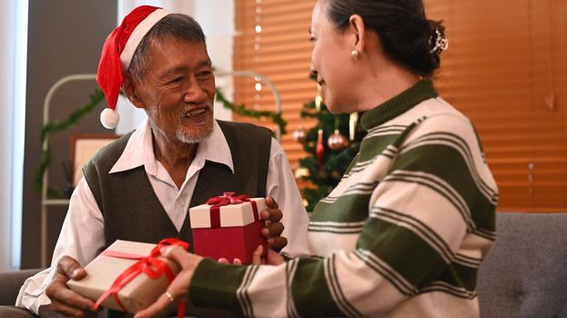 Photo of romantic senior couple sitting in front of Christmas tree and exchanging presents. Celebration, holidays and people concept.