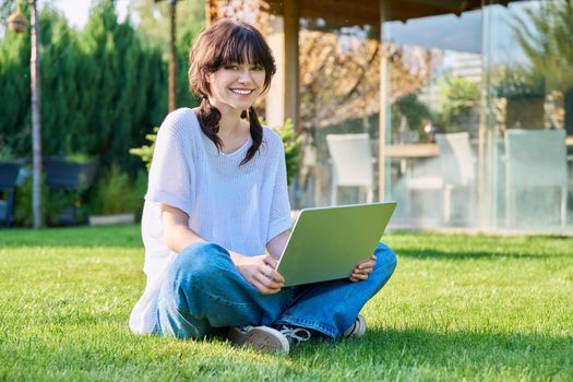 Teenage girl sitting on grass using laptop. Female teenager student 18, 19 years old sitting on lawn, looking at camera, technologies for studying leisure communication shopping in backyard in garden
