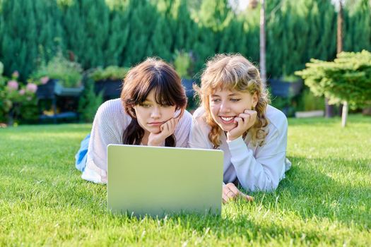 Two teenage girlfriends 17, 18 years old using laptop lying outdoors on grass on sunny summer day. Young female student looking at laptop screen, technology for leisure study entertainment