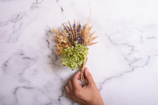 A bouquet of dry flowers and herbs in a woman's hand on a marble background.
