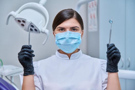Headshot portrait of female dentist wearing facial mask with instruments for examining teeth. Doctor looking at camera, posing in dental office. Dentistry, medicine, treatment, dental health care