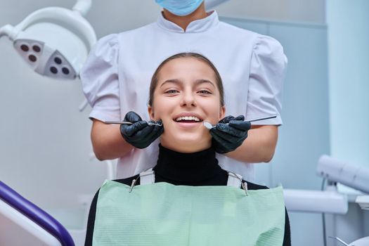 Portrait of young teenage girl in dental chair with hands of doctor with tools. Female teenager smiling with teeth looking at camera in dentist office. Adolescence hygiene treatment dental health care