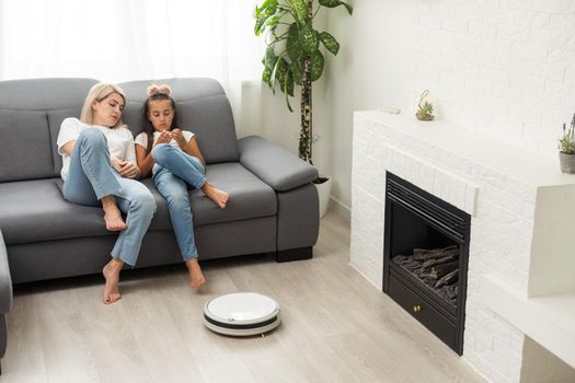 Cute child near mother looking at robotic vacuum cleaner in living room.