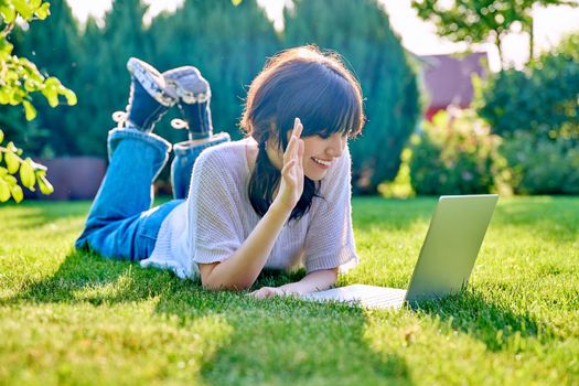 Young teenage female lying outdoor on the grass with a laptop talking with friends relatives via video call. Happy girl 18, 19 years old relaxing talking online using webcam at conference