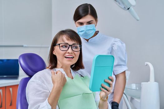 Happy middle aged woman together with dentist, patient sitting in dental chair looking at mirror. Prosthetics, treatment, implantation, dental teeth health, beauty care concept