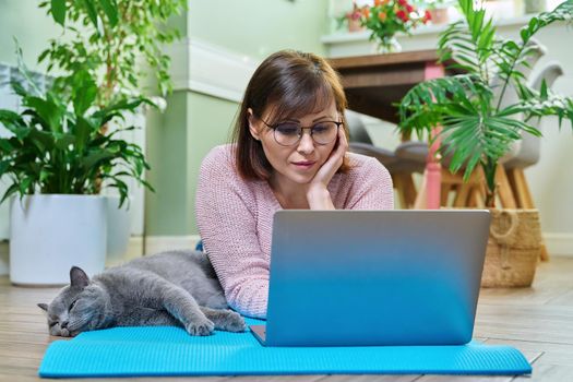 Middle-aged woman lying with cat on exercise mat, looking at laptop screen. Home leisure, lifestyle, activity, relaxation, people and pets concept
