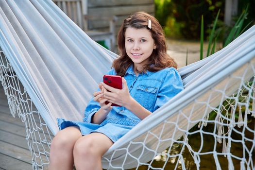 Outdoor portrait of teenage girl 12, 13 years old sitting in hammock with smartphone. Smiling girl looking at camera relaxing in backyard using mobile apps for study and leisure