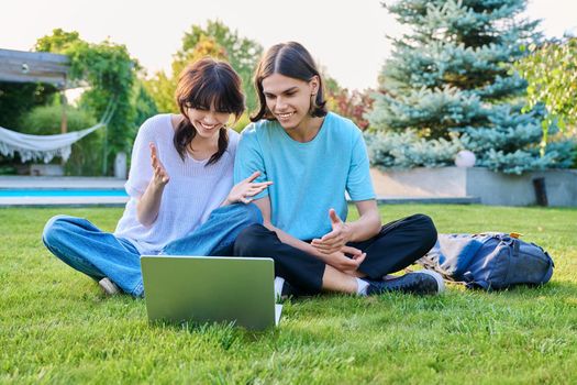 Two teenage friends of students sitting on grass with laptop, in backyard, guy and girl 18 years old study together. Friendship, youth, technology, high school, college, lifestyle concept