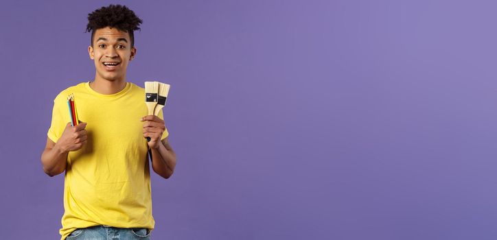 Portrait of upbeat young creative man, artist starting art courses, want to learn how to draw, holding colored pencils and brushes, standing enthusiastic over purple background.