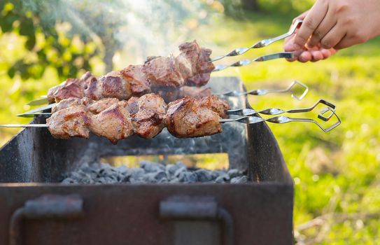 Cooking shish kebab on the grill with smoke, the man flips the skewers. Fresh meat for barbecue grilled on fire. Picnic in nature