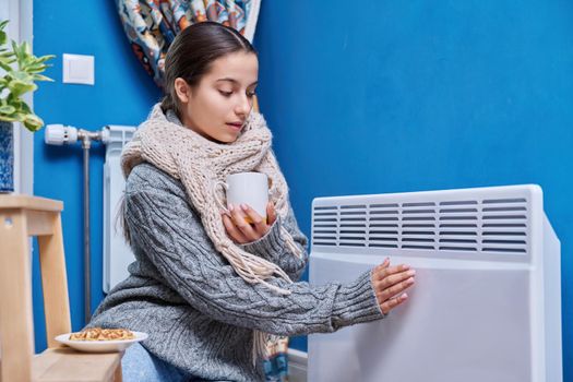 Cold autumn winter season, lifestyle, teenage female in warm sweater scarf warming near electric heating radiator, drinking hot drink with cup. Heating, warmth, equipment, life in cold season