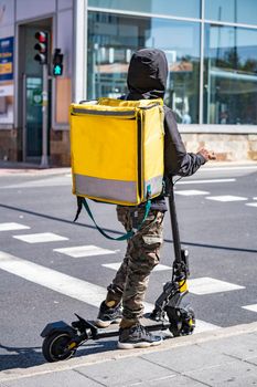Man courier food delivery with yellow thermal backpack waiting the green traffic light on an electric scooter.