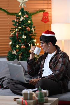 Young asian man in Santa hat drinking hot beverage and surfing internet with laptop at home.