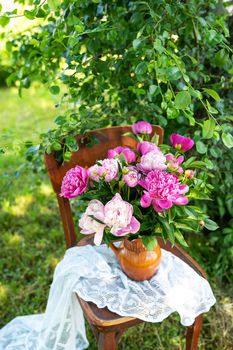 A large bouquet of pink peonies stand in a vase on a wooden old chair, a bridal bouquet, white fabric