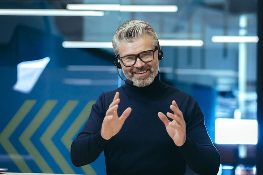 Senior gray haired businessman talking on video call, using headset investor in glasses smiling and looking at camera, webcam view man working inside modern office building.