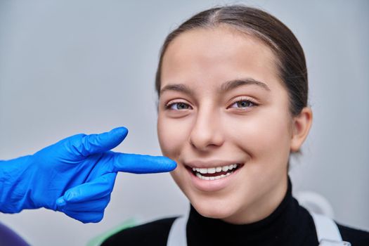 Close-up face smiling young teenage female, dentist hands showing healthy beautiful teeth with luxury jewelry rhinestone dental diamond on patient tooth. Beauty, dentistry, fashion concept