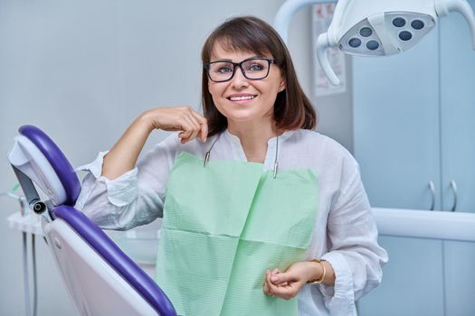 Happy middle aged woman patient looking at camera sitting in dental chair in dentist office. Smiling female after taking care of teeth whitening treatment, dental implant prosthetics