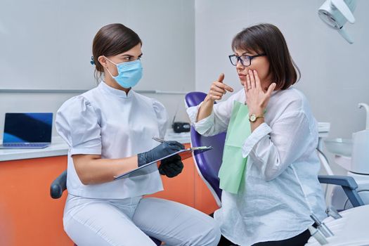 Dentist and woman patient in dental clinic, doctor nurse writing on clipboard consulting, middle aged female talking about toothache disturbing dental problem. Dentistry, treatment, dental health care