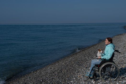 A serene Caucasian woman in a wheelchair is resting on the seashore with a jack russell terrier dog