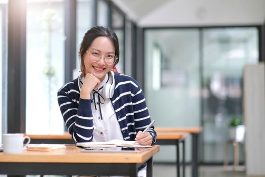 Asian woman wearing headphones using laptop in cafe, writing notes, attractive female student learning language, watching online webinar, listening audio course, e-learning education concept..