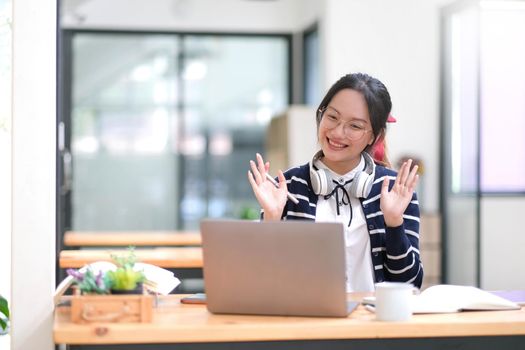 Smiling asian woman waving hello at video chat on laptop, sitting at home, studying remotely in e-learning school.