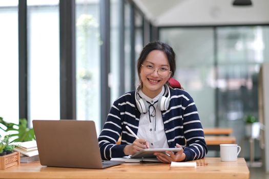 Young adult happy smiling Hispanic Asian student wearing headphones talking on online chat meeting using laptop in university campus or at virtual office. College female student learning remotely..