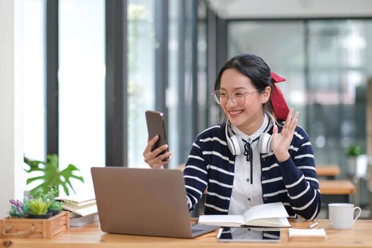 Female student taking notes from a book and using smart phone at cafe. Young asian woman sitting at table doing assignments in cafe..