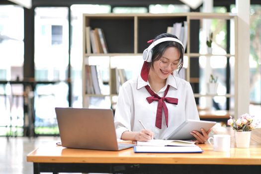 Happy young Asian student woman wearing headphones looking at webcam, looking at camera, during virtual meeting or video call talk..