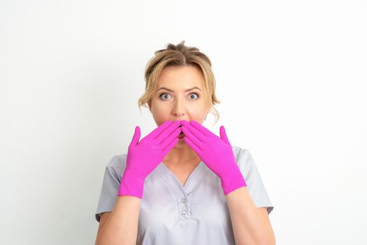 Portrait of a young female caucasian doctor or nurse is shocked covering her mouth with her pink gloved hands against a white background