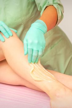 Sugaring legs. Woman legs hair removing. Hands in pink and blue rubber gloves of two beauticians apply sugar paste on female feet