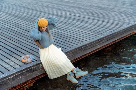 Outdoors fashion portrait of a beautiful middle aged woman walking on the beach. Marine background. Dressed in a stylish warm blue sweater, yellow skirt and beret