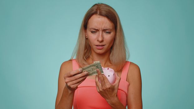 Poor woman upset over insufficient amount of money, holding piggybank and one dollar banknote. Financial crisis. Bankruptcy. Poverty and destitution. Young girl isolated on blue studio wall background