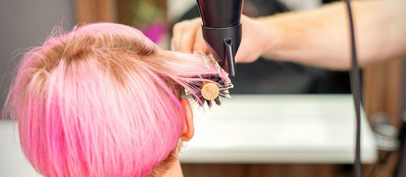 A professional hairdresser is drying long red hair with a hair dryer and round brush, close up