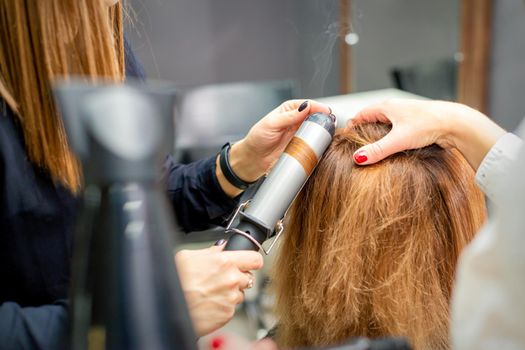 Hands of female hairstylist curls hair client with a curling iron in a hairdressing salon, close up