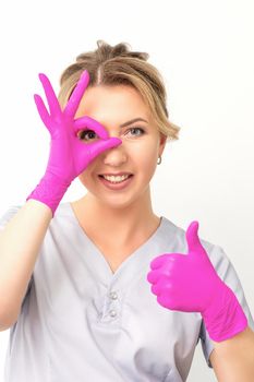 Smiling doctor oculist caucasian woman wearing pink rubber gloves in uniform showing ok sign covering the eye and thumb up gesture against a white background