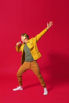 Singing young man enjoying song dancing with one hand up using phone and wireless headphones wearing jeans yellow jacket isolated on red background. Man sing while recording his voice.