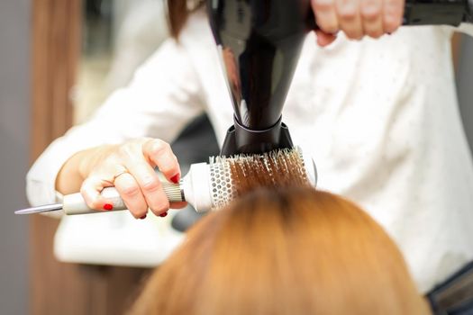 A professional hairdresser is drying long red hair with a hair dryer and round brush, close up