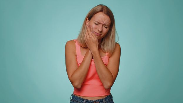 Blonde young woman touching sore cheek suffering from toothache cavities or gingivitis waiting for dentist appointment, gums disease. Adult girl indoors studio shot isolated alone on blue background