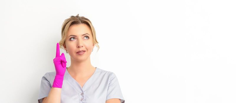 Portrait of young caucasian doctor woman wearing rubber gloves in medical uniform with one finger up, looking up against a white background, copy space