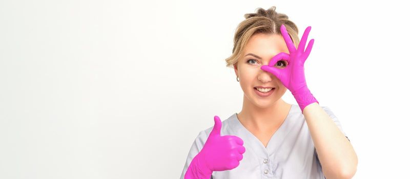 Smiling doctor oculist caucasian woman wearing pink rubber gloves in uniform showing ok sign covering the eye and thumb up gesture against a white background