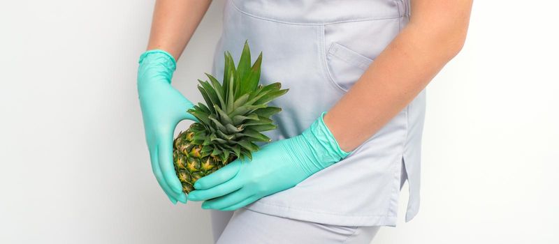 Young beautician wearing blue gloves in uniform with pineapple covers an intimate area on a white background, bikini zone depilation concept