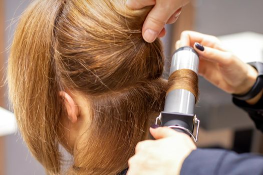 Hands of female hairstylist curls hair client with a curling iron in a hairdressing salon, close up