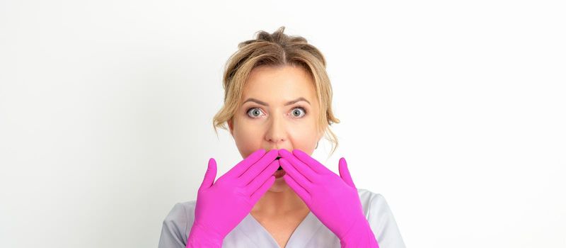 Portrait of a young female caucasian doctor or nurse is shocked covering her mouth with her pink gloved hands against a white background