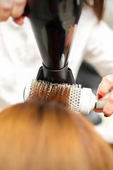 A professional hairdresser is drying long red hair with a hair dryer and round brush, close up