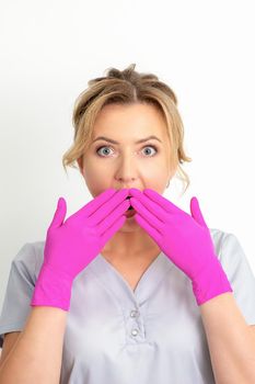 Portrait of a young female caucasian doctor or nurse is shocked covering her mouth with her pink gloved hands against a white background