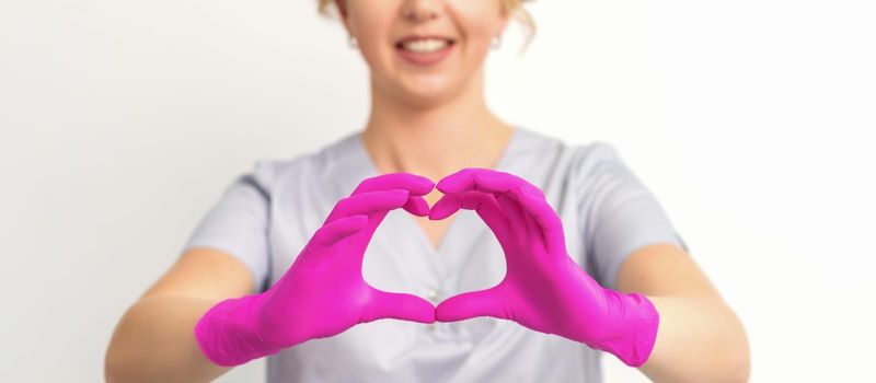 A smiling caucasian woman doctor wearing pink gloves in uniform showing the symbol of a heart against a white background